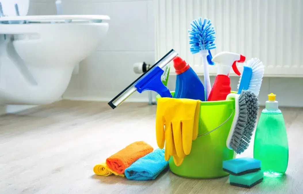 A bucket filled with various bathroom cleaning supplies, including brushes, gloves, spray bottles and sponges, placed on a wooden floor near a toilet.