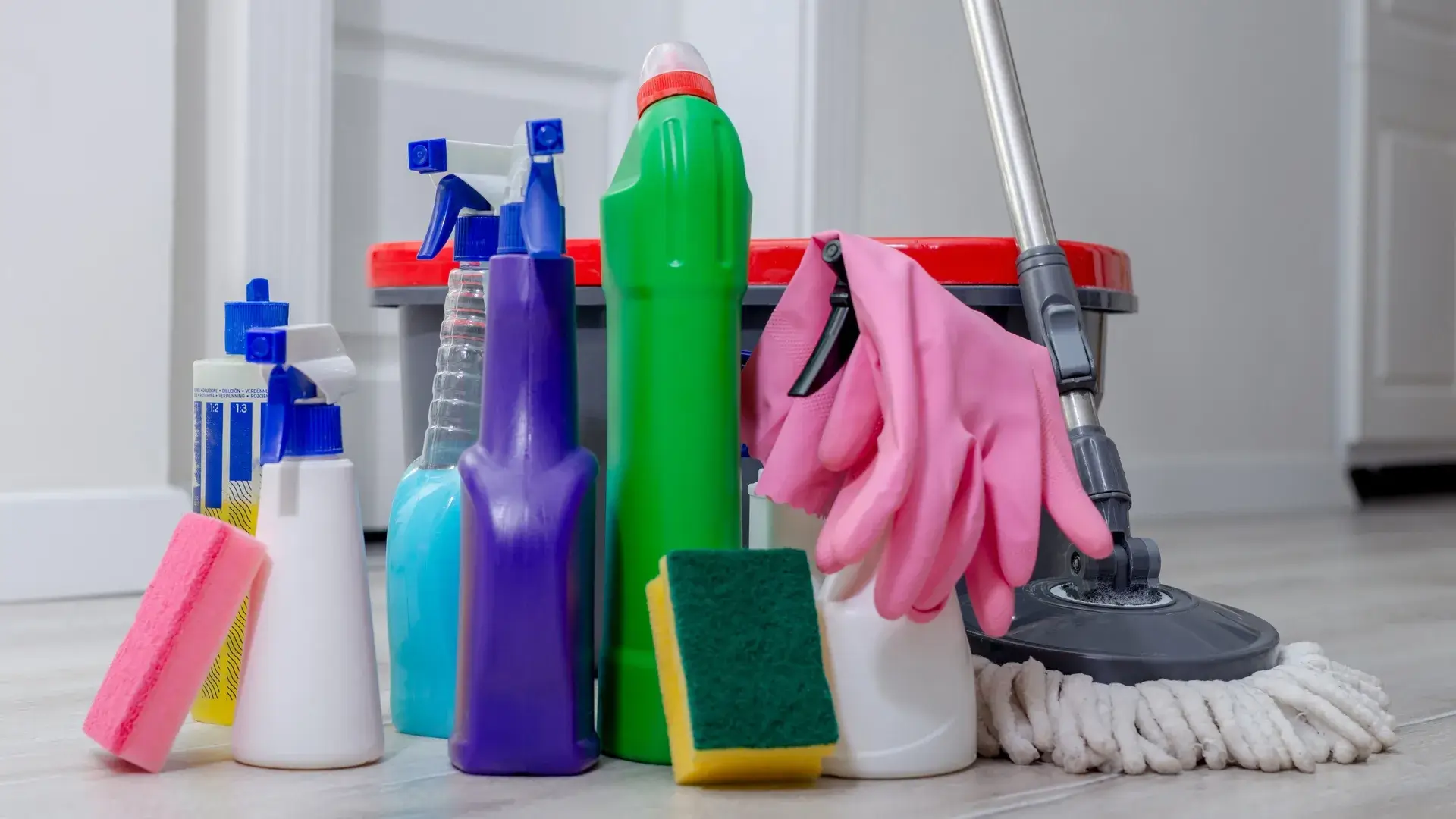 A collection of cleaning supplies, including spray bottles, sponges, gloves and a mop bucket, arranged on a clean floor.