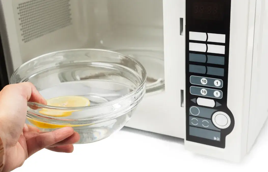 Hand placing a bowl of water with lemon slices into a microwave for cleaning.