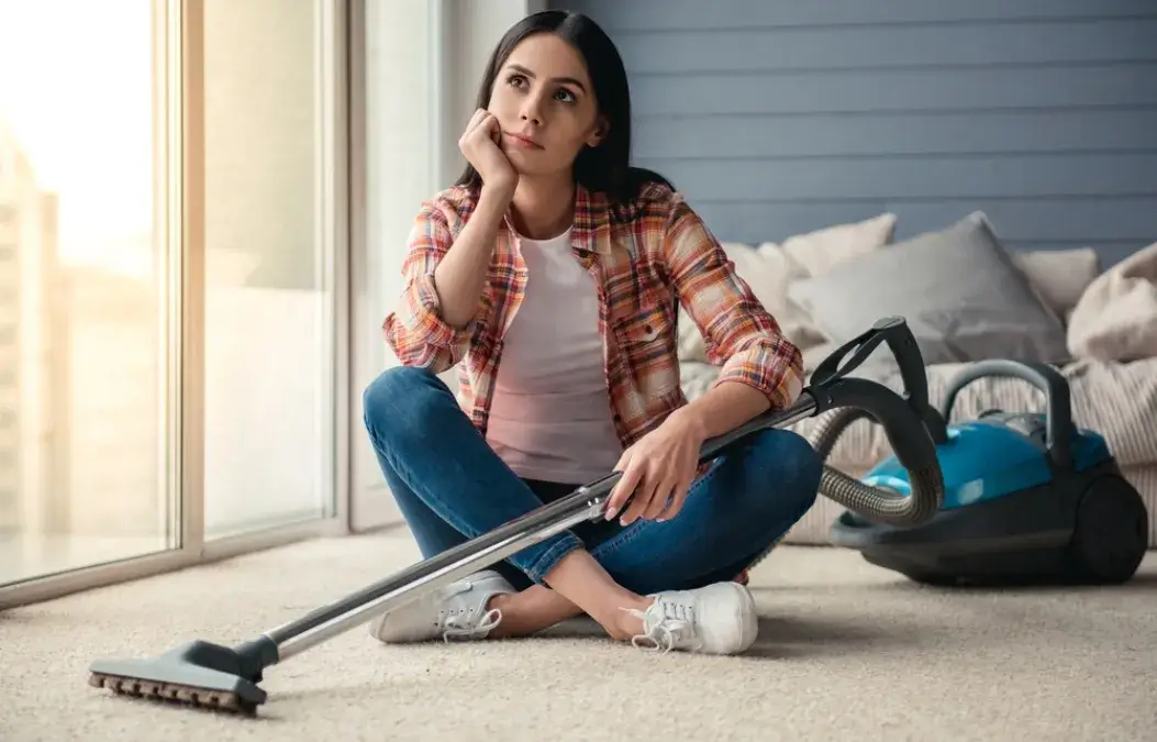 Young woman sitting on the floor with a vacuum cleaner, looking thoughtful while taking a break from cleaning.