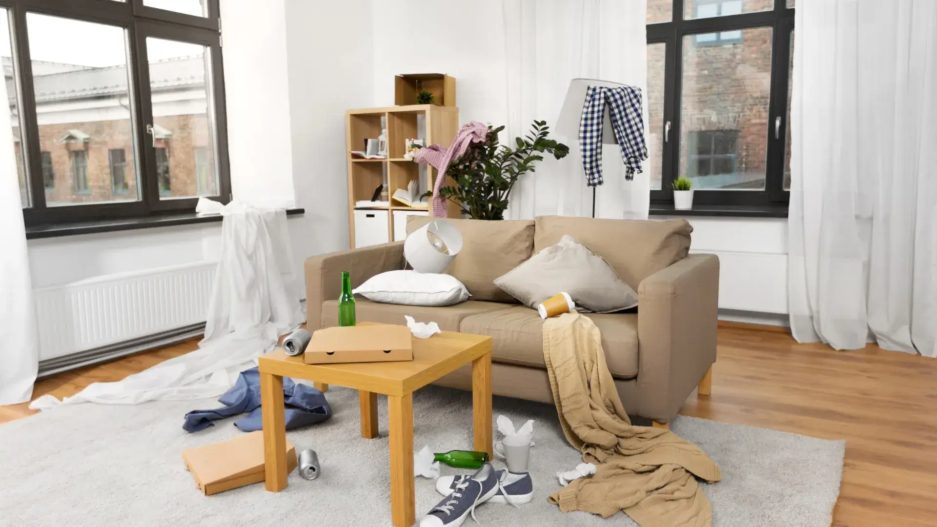 A messy living room with scattered clothes, empty bottles and trash on the floor and couch.