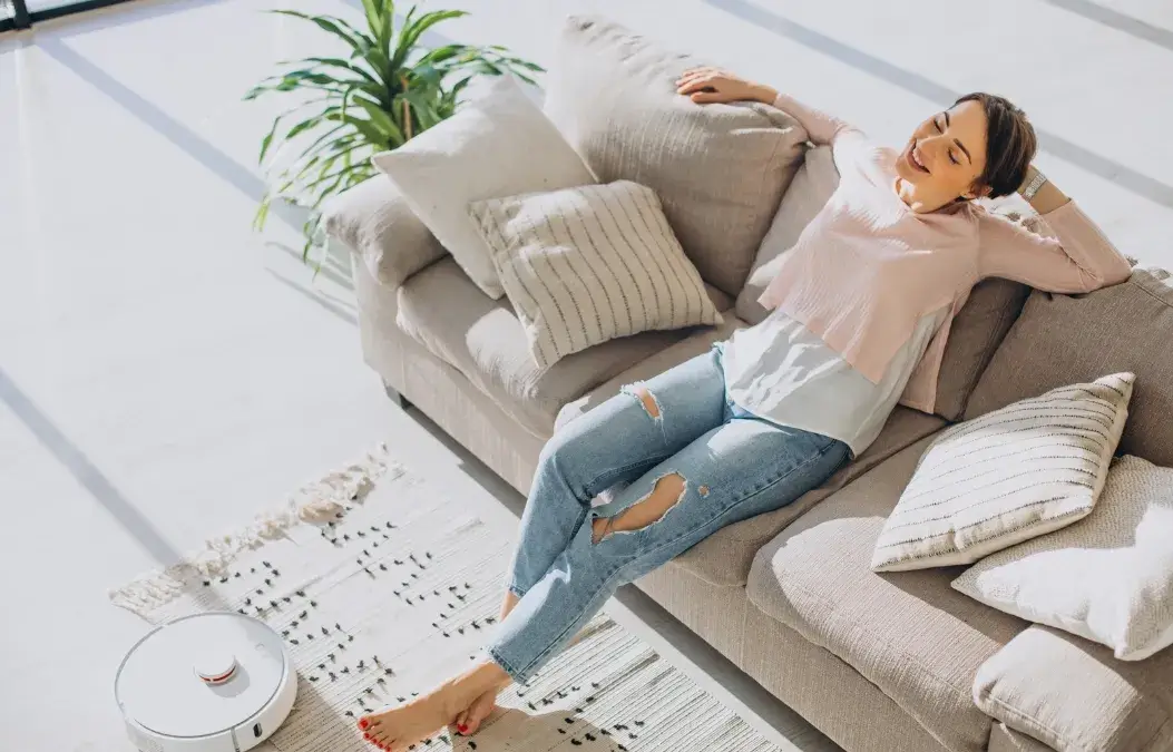 A woman relaxing on a beige couch in a bright living room with a robotic vacuum nearby.