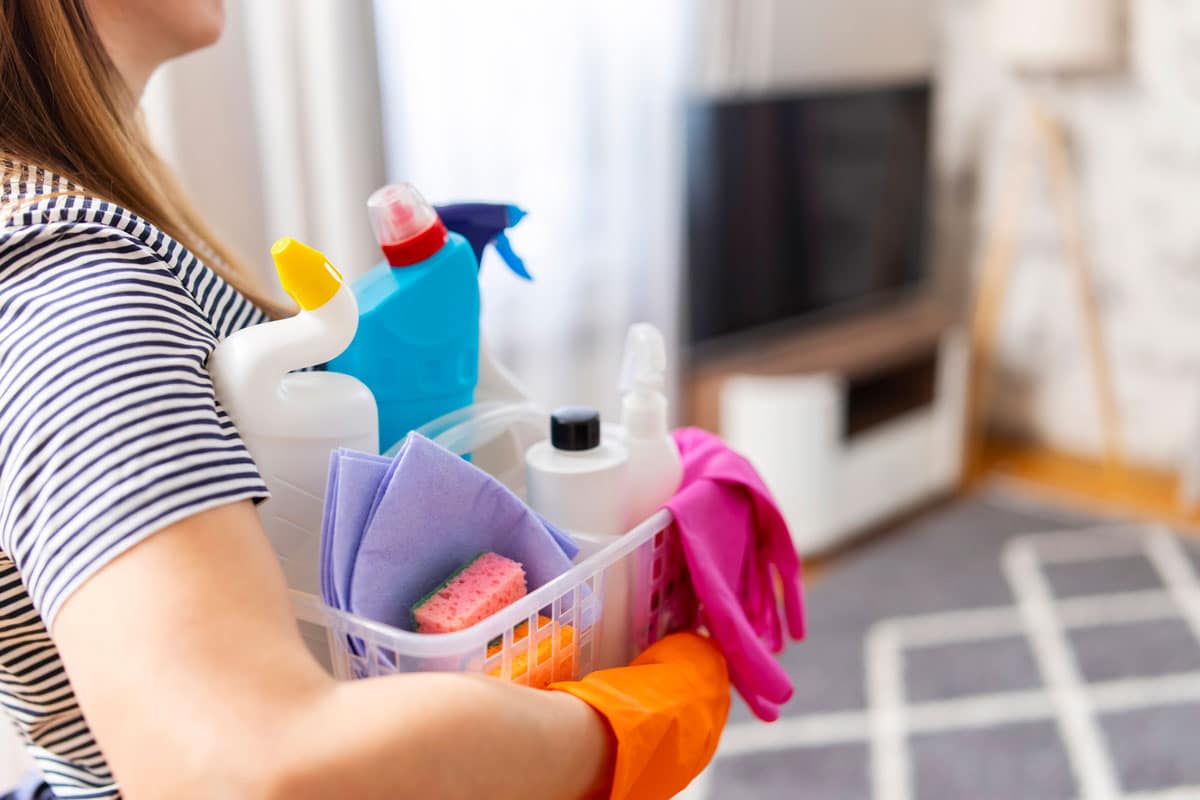 Woman holding a plastic caddy filled with colorful detergents and microfiber cloths for a regular cleaning services in a living room.