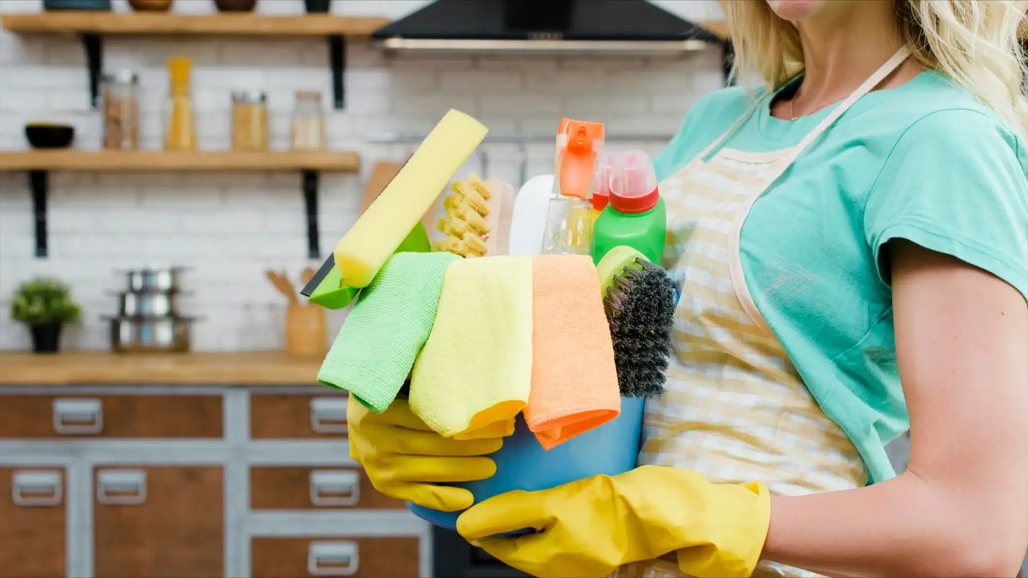 Woman in yellow gloves holding a blue bucket of cleaning products in a kitchen.