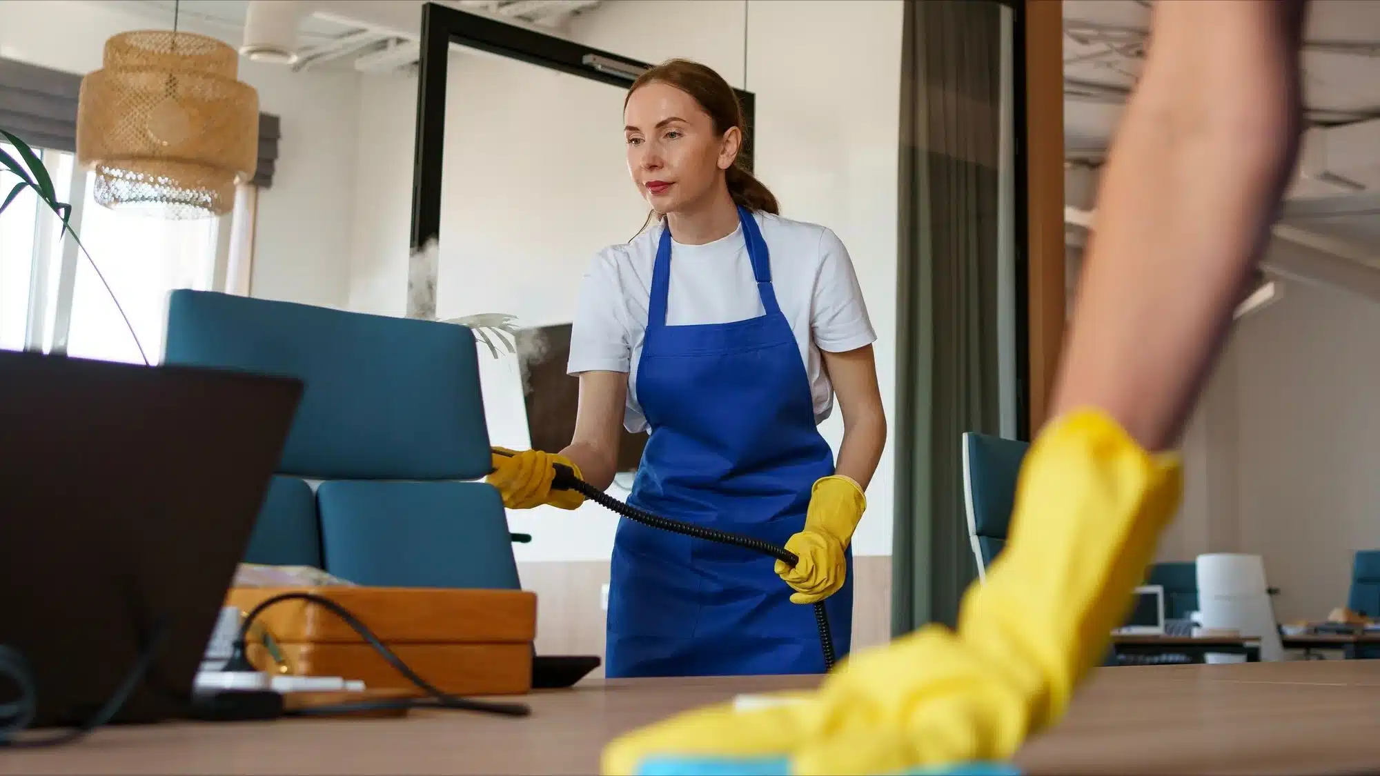 Woman in a blue apron steam cleaning an office desk.