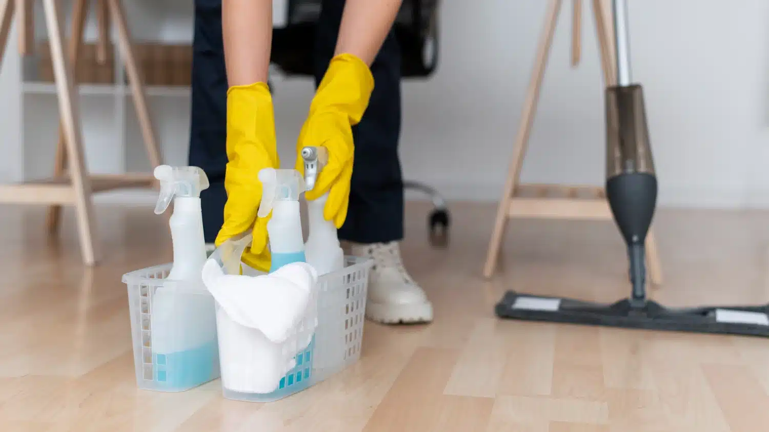 Cleaner in yellow gloves picking up a basket of supplies from an office floor.