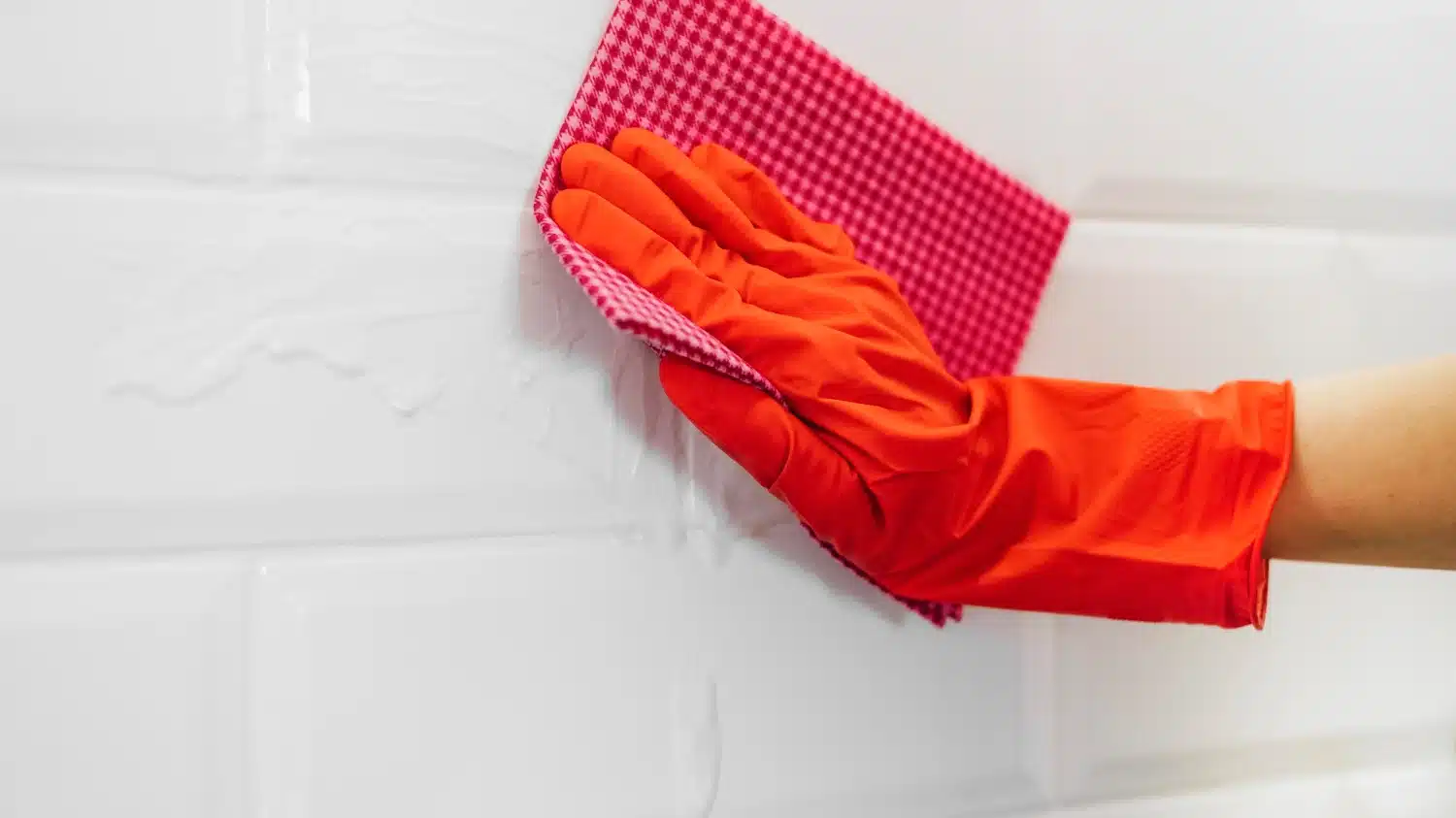 Red-gloved hand wiping white subway tiles with a damp cloth.