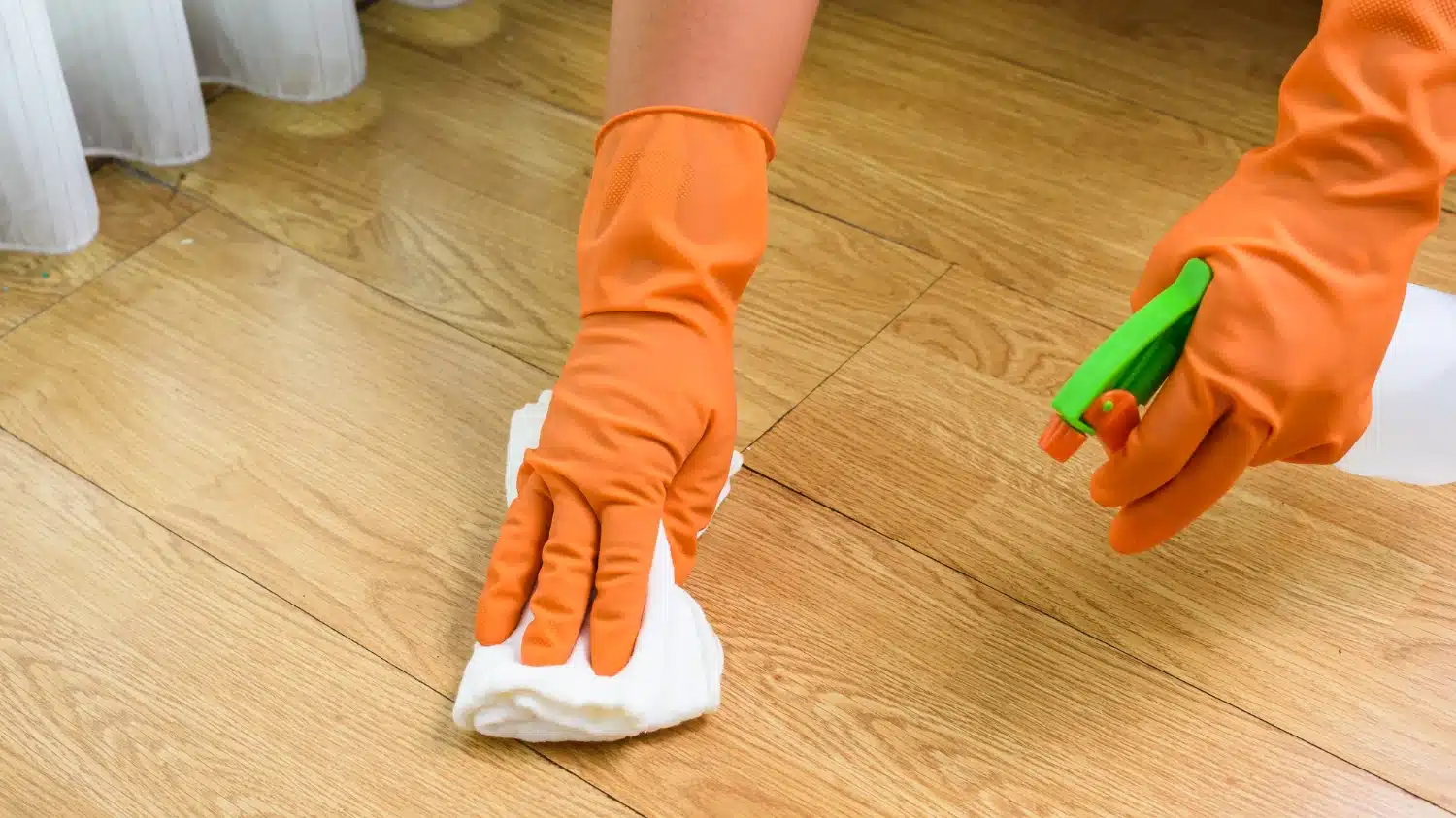 Orange-gloved hands scrubbing a hardwood floor with a cloth and spray bottle.