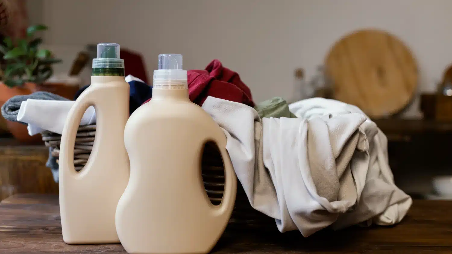 Two detergent bottles beside a basket of mixed laundry on a wooden surface.