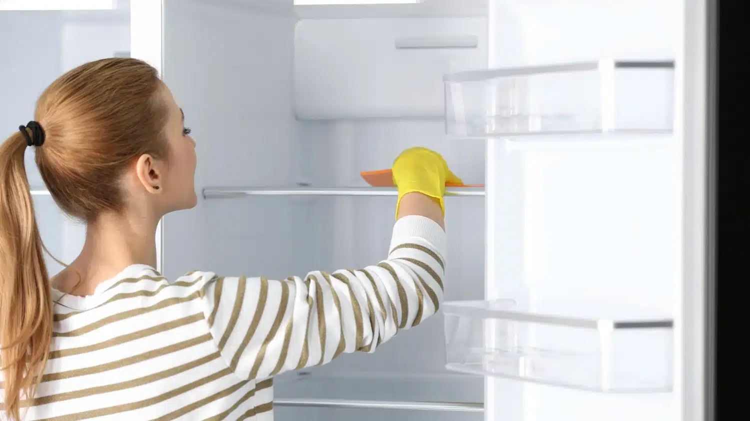 Woman wiping empty refrigerator shelves with a yellow glove.