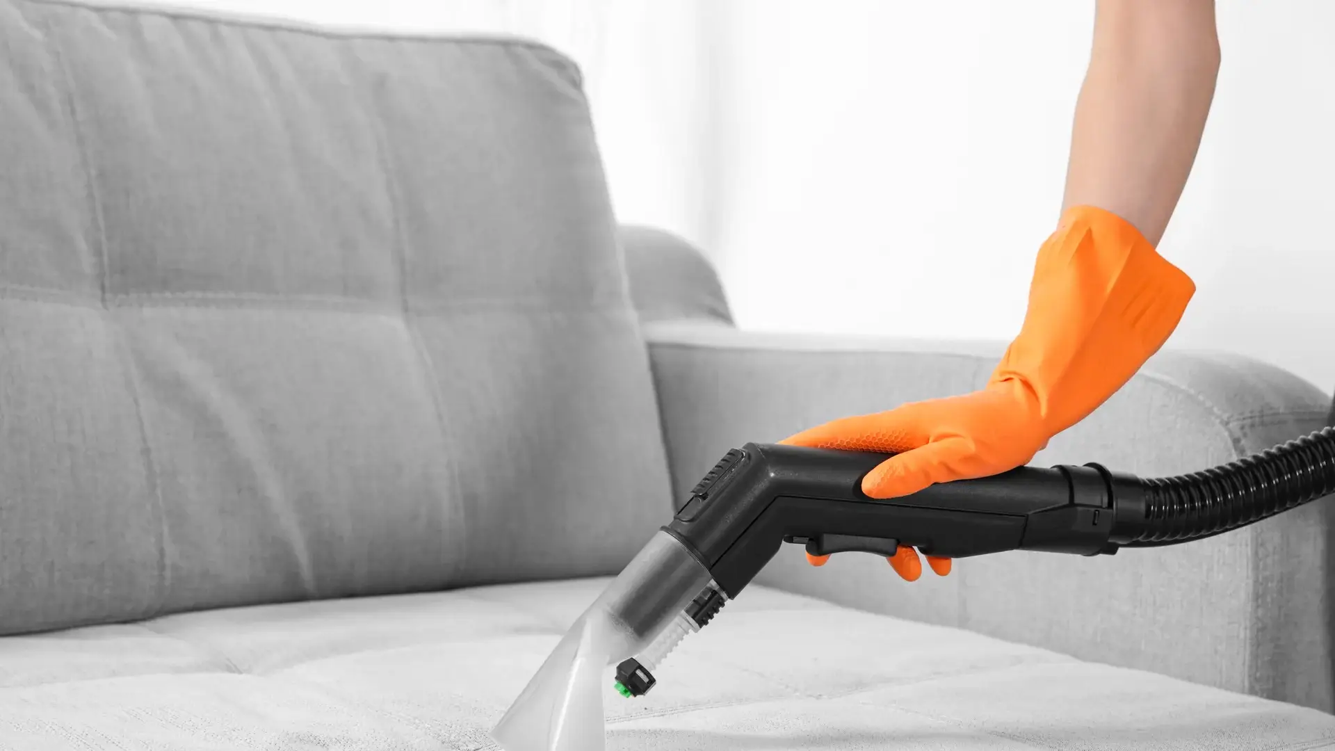 Hand with orange glove using vacuum cleaner on gray sofa
