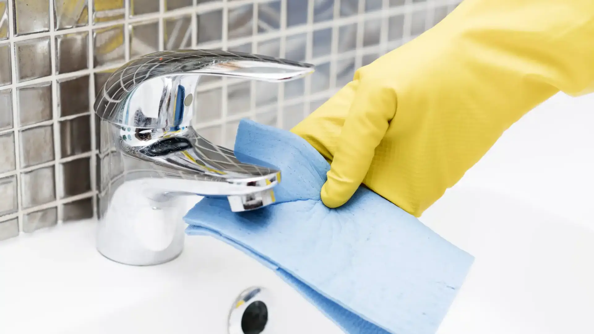 Yellow gloved hand cleaning a chrome bathroom faucet with a blue cloth