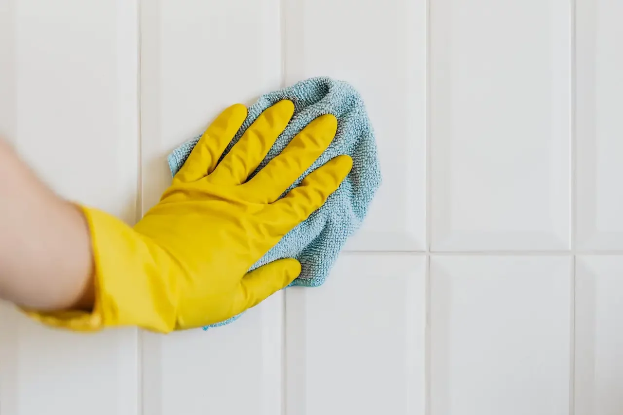 Gloved hand cleaning a bright white tile wall with a blue microfiber cloth