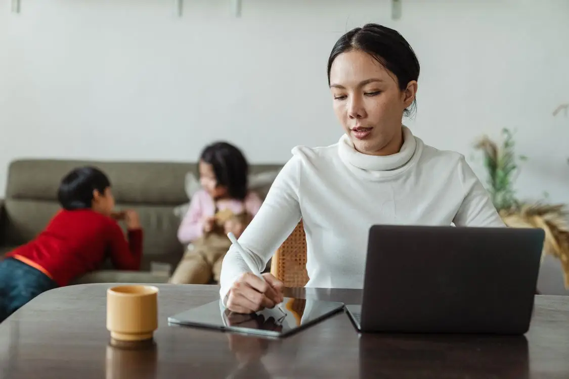 A parent multitasking on a laptop at the kitchen table while kids play in a bright, tidy Frisco home